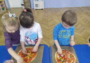 Troje dzieci układa na pizzy poszczególne skladniki.